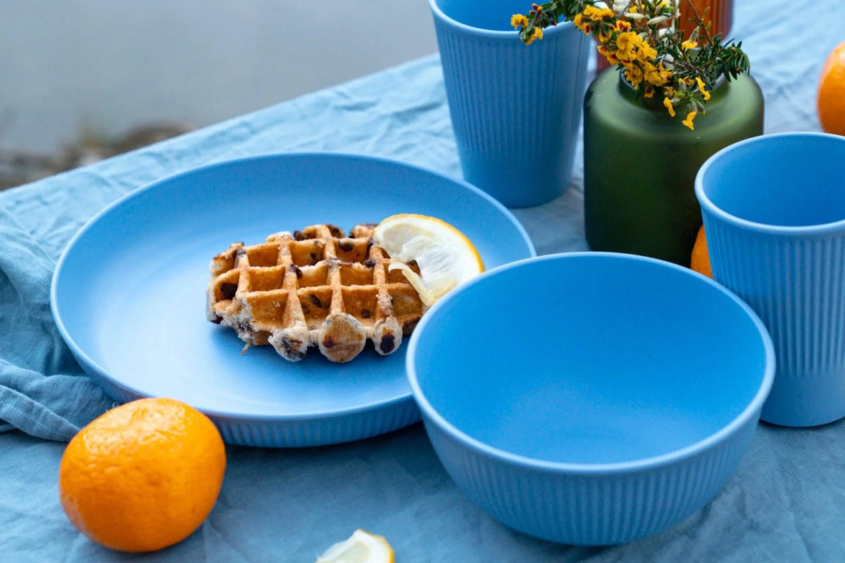 A set of bright blue ceramic tableware, including a plate, bowl, and cups, styled on a blue fabric tablecloth with a waffle, orange, and a green vase in the background.