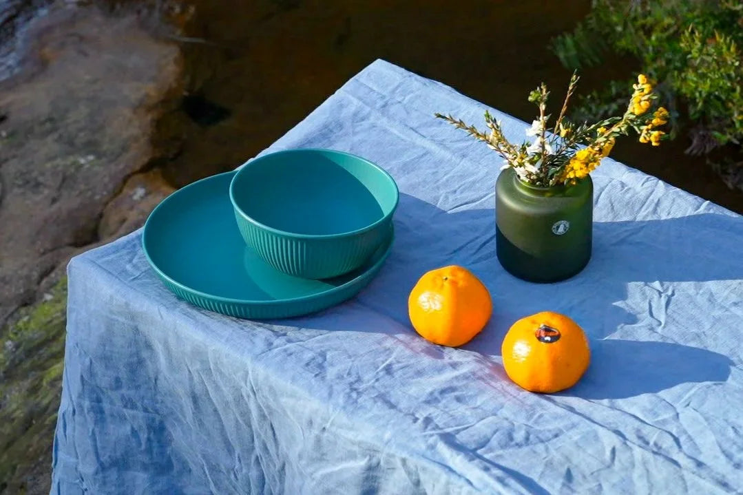 A teal green ceramic bowl and matching tray set on a light blue tablecloth outdoors, styled with two oranges and a dark green glass vase holding yellow flowers.