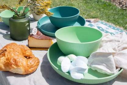 A styled outdoor picnic scene featuring a set of pastel green and teal ribbed bowls and plates. The table setting includes a golden croissant, a slice of cheesecake, white marshmallows, a green ceramic vase with dried yellow flowers, and a white cloth, all set against a blurred background of green grass.
