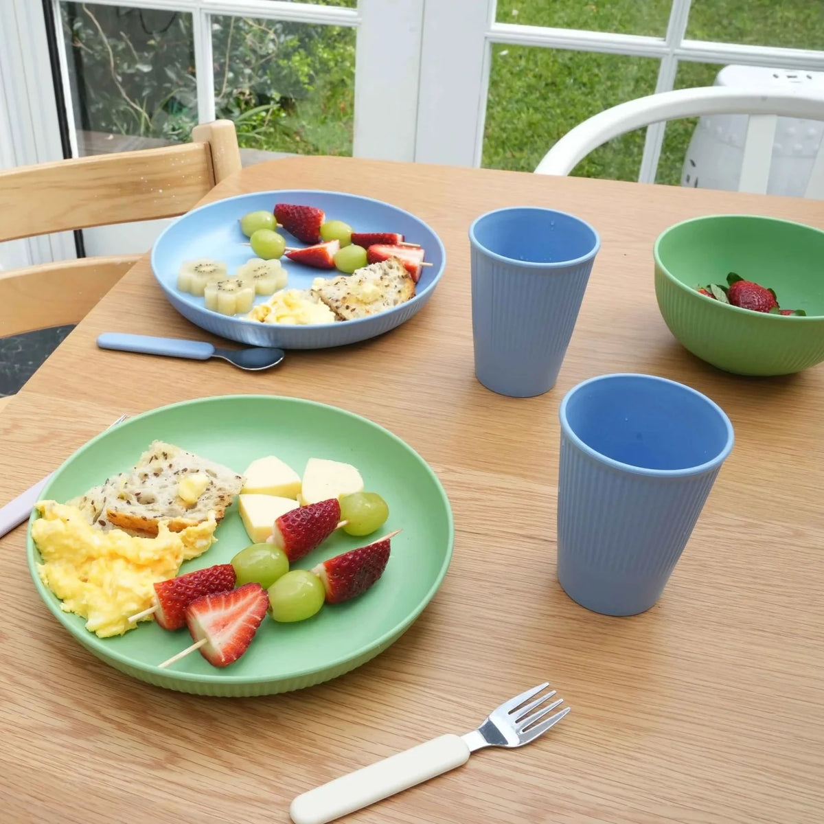 A children's breakfast table setting featuring a light green plate with scrambled eggs, cheese, and fruit skewers, a blue plate with sliced fruit and omelet, two ribbed blue cups, a green bowl of strawberries, and a fork on a wooden table.