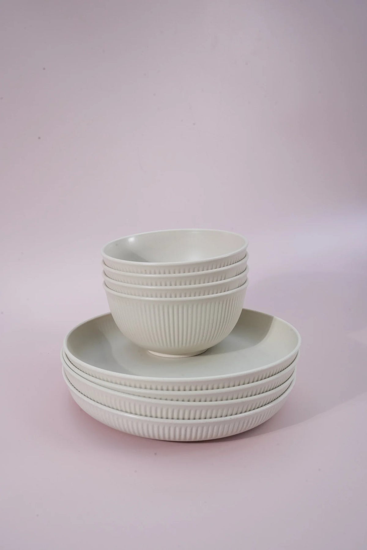 A set of white ribbed ceramic bowls and plates stacked neatly on a soft pink studio background.