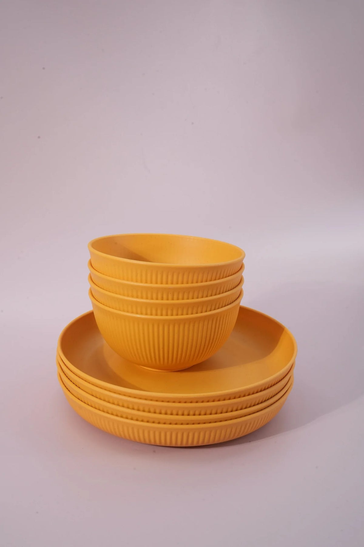 A set of mustard yellow plastic tableware, including a stack of four ribbed bowls and a matching stack of four ribbed plates, photographed on a light gray studio background.