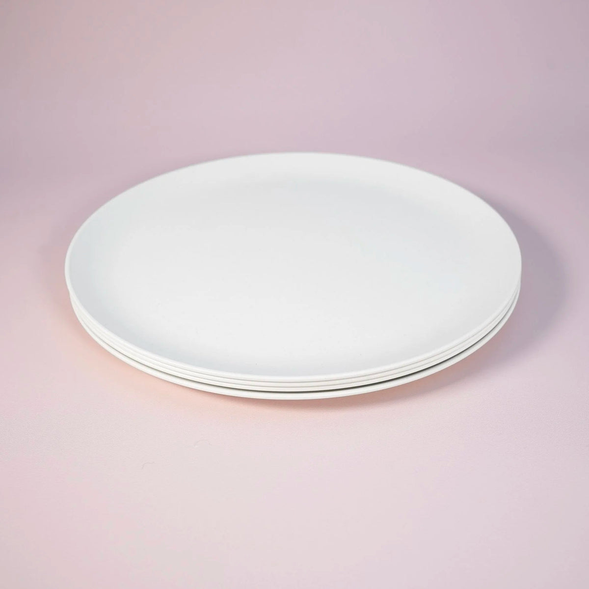 A stack of four plain white ceramic plates photographed on a soft pink studio background.