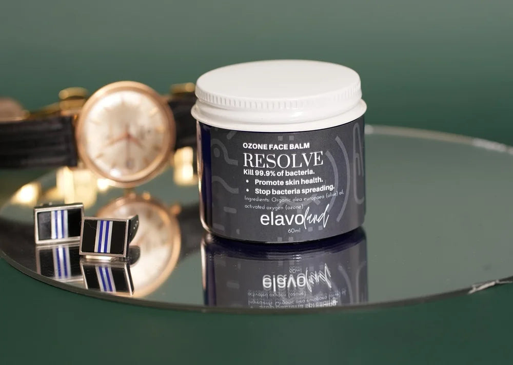 A jar of elavocean Resolve Ozone Face Balm sits on a reflective dark green surface, styled with a gold watch and striped cufflinks in the background.