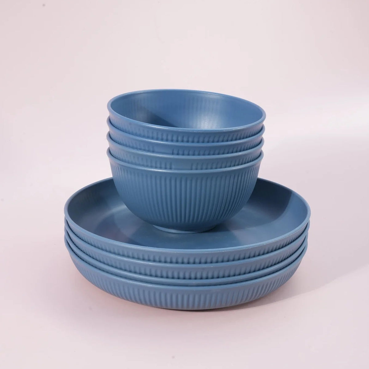 A set of blue ribbed ceramic dinnerware, including a stack of bowls and matching plates, photographed on a clean, light pink studio background.