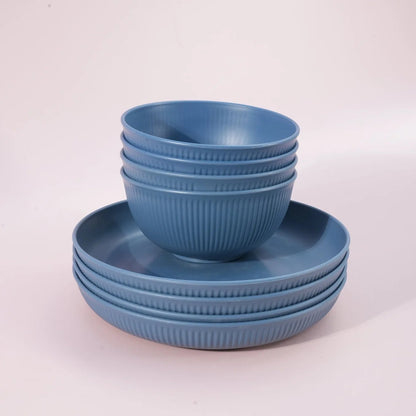A set of blue ribbed ceramic dinnerware, including a stack of bowls and matching plates, photographed on a clean, light pink studio background.