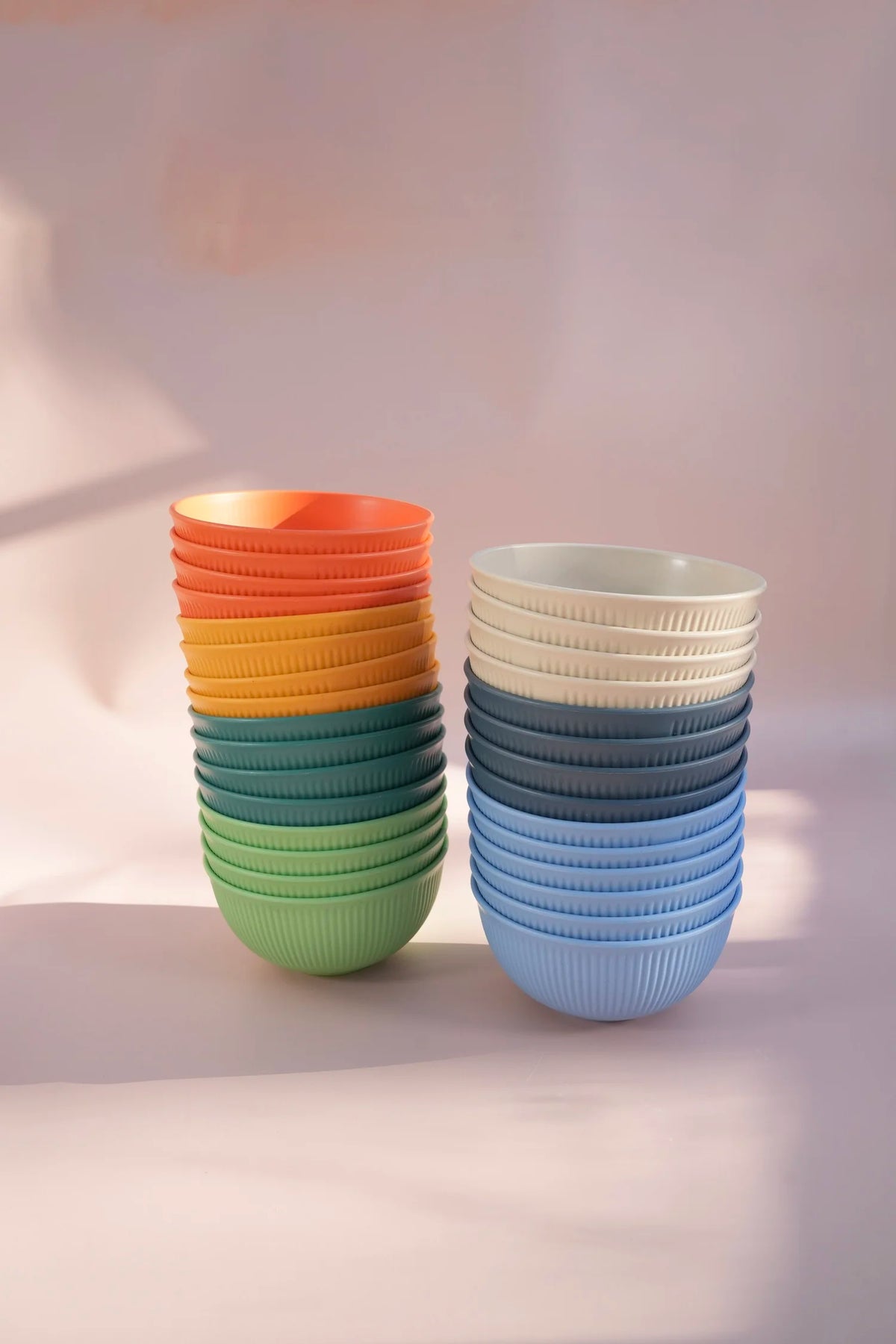 Two stacks of ribbed ceramic bowls in gradient colors, one showing orange, yellow, teal, and green, and the other showing off-white, dark blue, and light blue, set against a soft pink background.