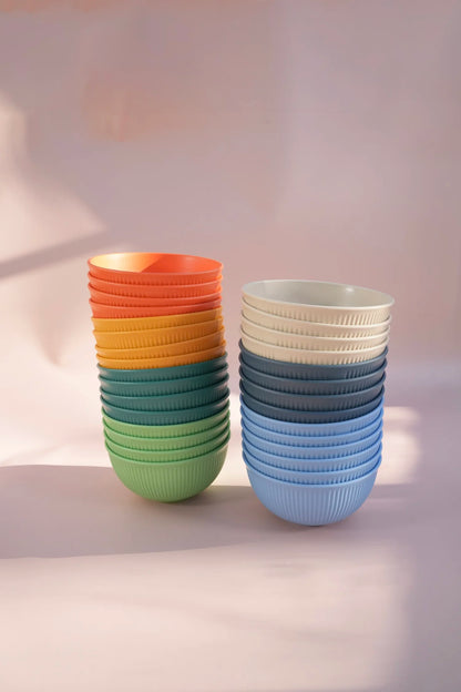 Two stacks of ribbed ceramic bowls in gradient colors, one showing orange, yellow, teal, and green, and the other showing off-white, dark blue, and light blue, set against a soft pink background.