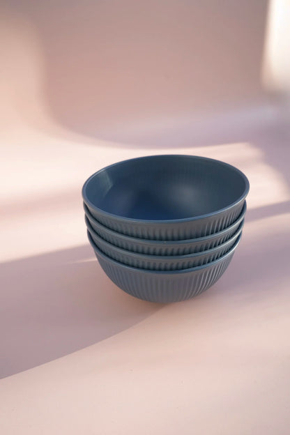 A stack of four ribbed ceramic bowls in a muted slate blue color, set against a warm, softly lit beige background.