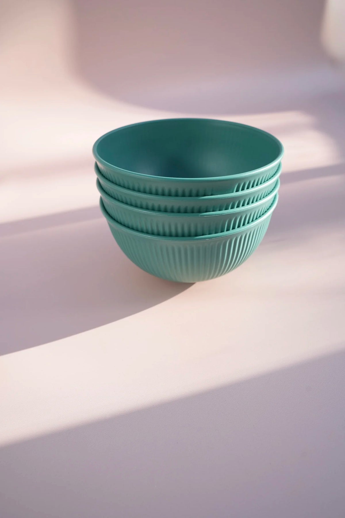 A stack of four ribbed ceramic bowls in a muted teal-green color, set against a soft, light pinkish-beige background with artistic diagonal shadows.