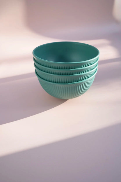 A stack of four ribbed ceramic bowls in a muted teal-green color, set against a soft, light pinkish-beige background with artistic diagonal shadows.