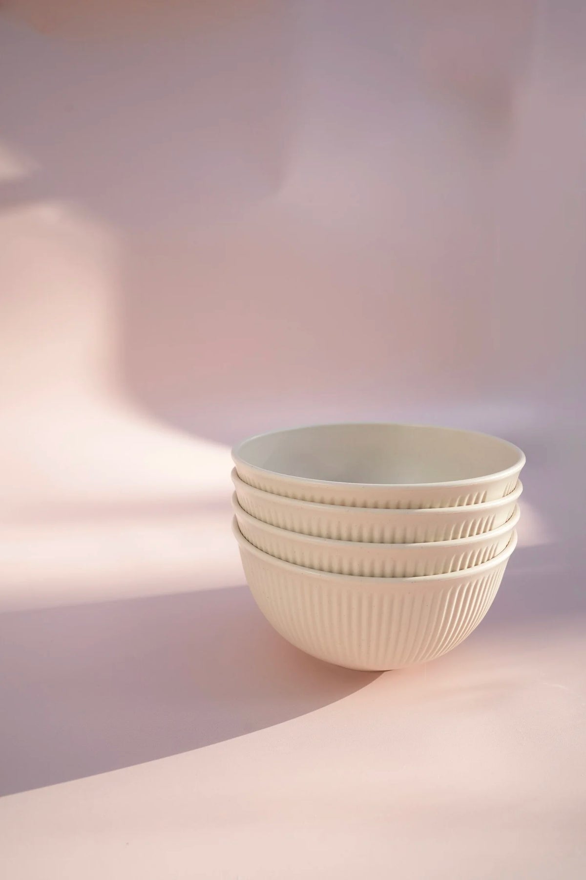 A stack of four ribbed ceramic bowls in a warm off-white color, set against a soft pink background with artistic shadows.