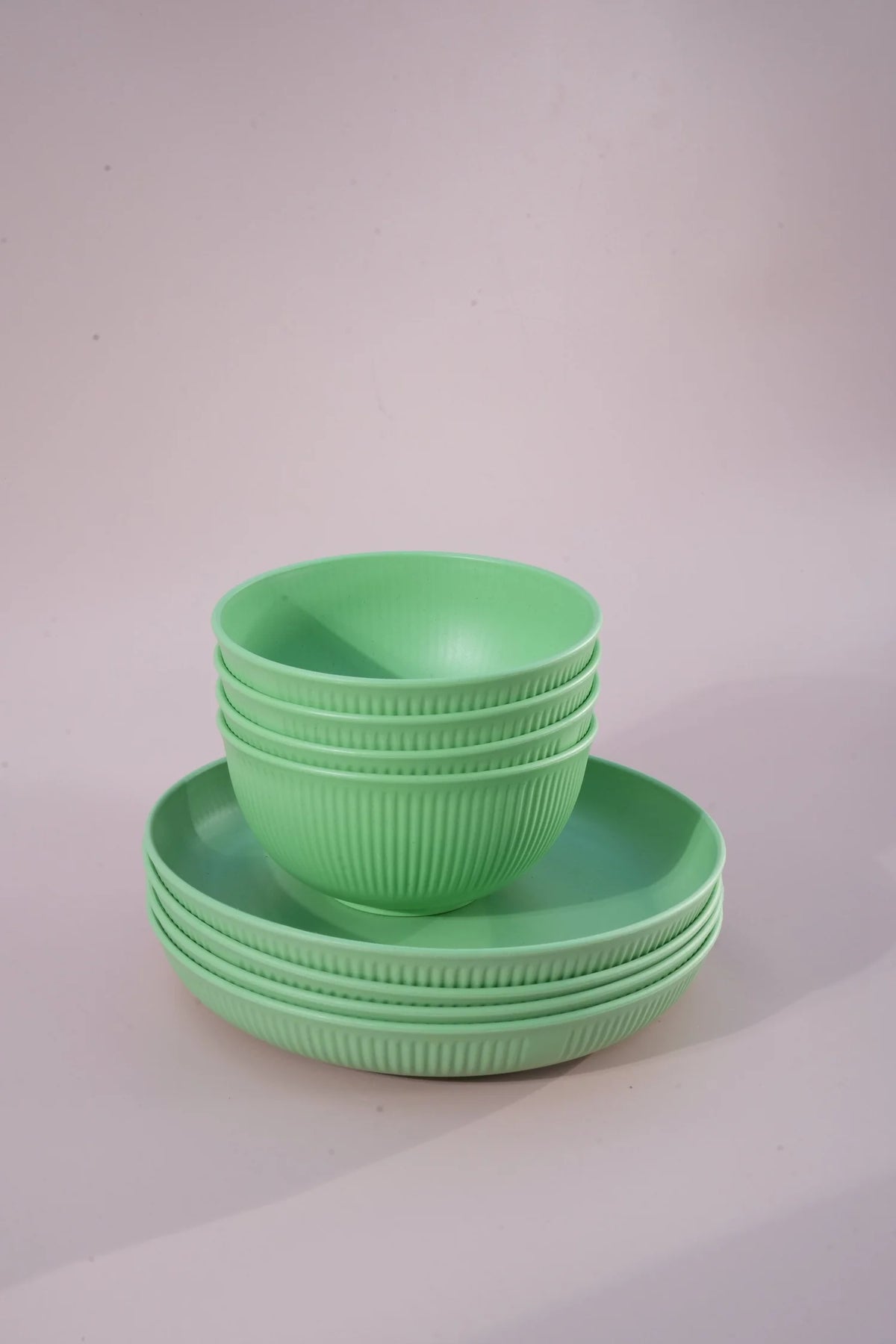 A set of six light green plastic bowls and plates with a ribbed exterior, stacked neatly on a light pink studio background.