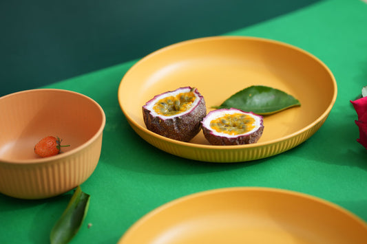 A yellow plate holding two halves of a passion fruit with a green leaf, styled on a bright green table alongside matching peach bowls and additional fruit.