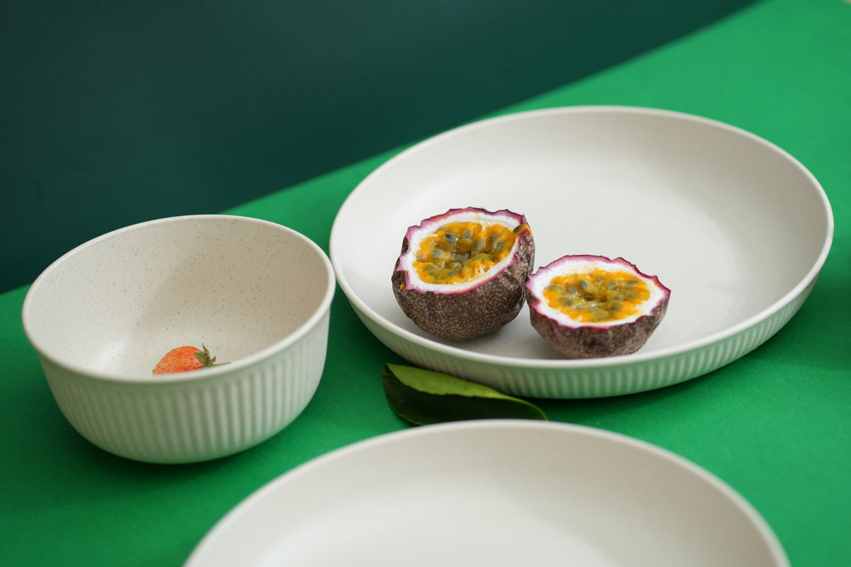 Three white ceramic bowls and plates on a green surface, with one plate holding two halves of a passion fruit and a small strawberry in the bowl.
